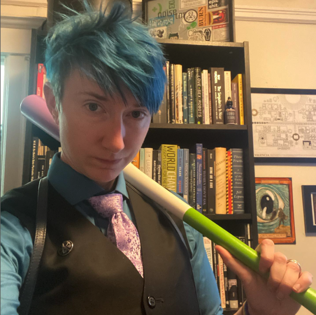 Willow stands in front of a book case and art in a vest, tie, and dress shirt. They hold a baseball bat painted in the genderqueer flag colors over one shoulder