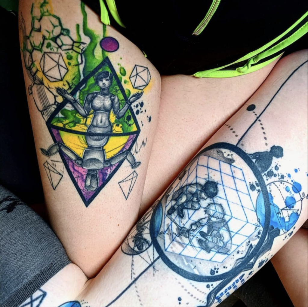 Tilde and Willow's right thighs are nestled together, with Tilde's tattoo of purples and greens with a mirrored person as posable figure on one side and a more realistic human on the other. Behind the realistic person, water color and shapes. Beyond the model, simpler shapes and more contained colors.On Willow's thigh, a circle surrounds two children poking at an ant hill. Outside the circle, a child's sillouette looks at plants in orbit. Another small circle holds an ant. There is blue and black water color around it all.
