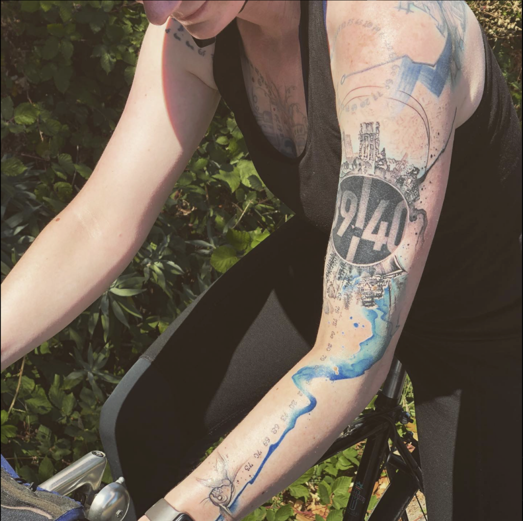 Willow rides a bike. Towards the top of their left arm is a circle with the numbers 39 40 on it, a city scape above it, and a forest with a ship below it. Blue water color streaks down the arm, with numbers alongside it, down to the wrist. At the wrist is a cute little fish.