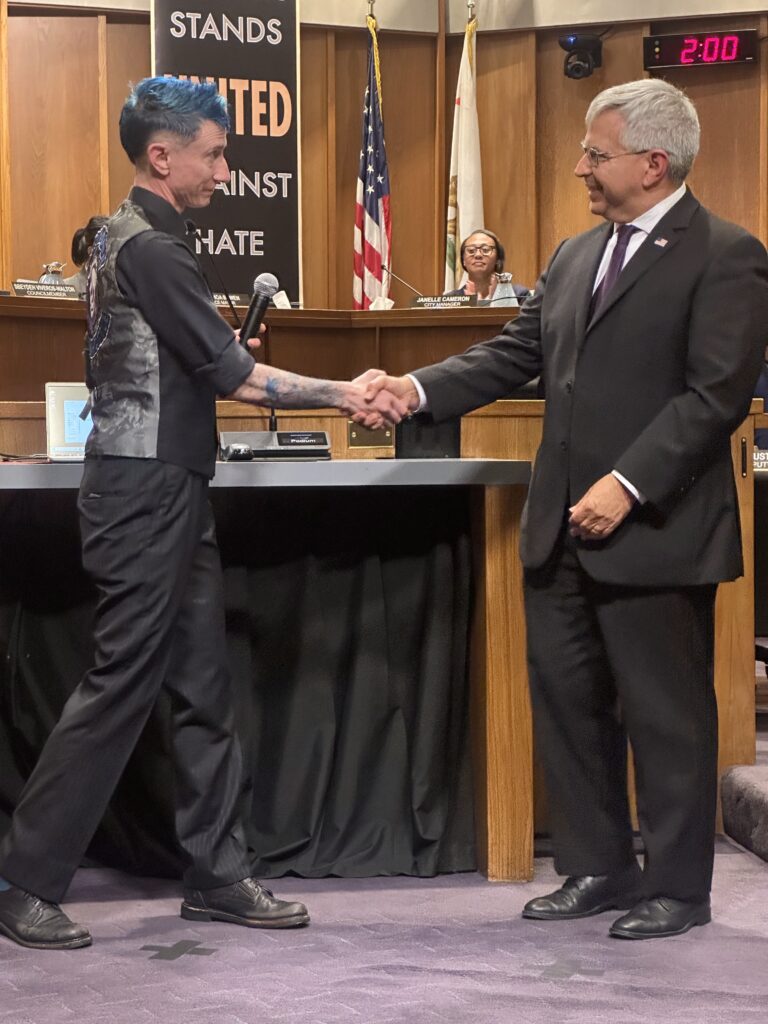 Willow shaking Mayor Gonzalez's hand in the city council chambers. They are both wearing suits and ties. Willow's hair is blue.