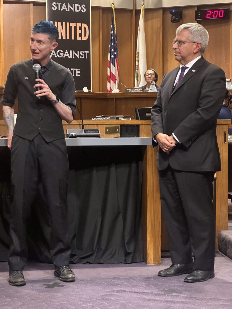 Willow giving a speech next to Mayor Gonzalez in the city council chambers. They are both wearing suits and ties. Willow's hair is blue and they wear a button that says "fix shit up."