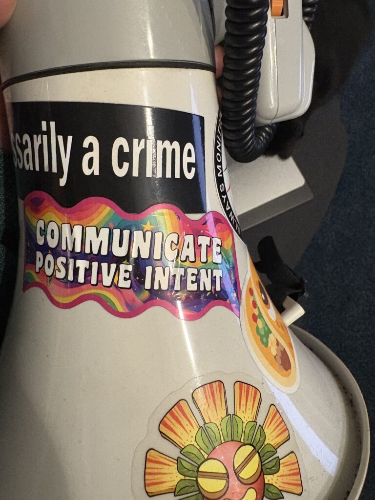 A picture of a megaphone focused on a sticker in Lisa Frank obnoxiously bright styling, "communicate positive intent." Additional stickers that are visible are the Priceless Baroot, the edge of a pleading taco emoji, and one that seems to ready "...necessarily a crime."