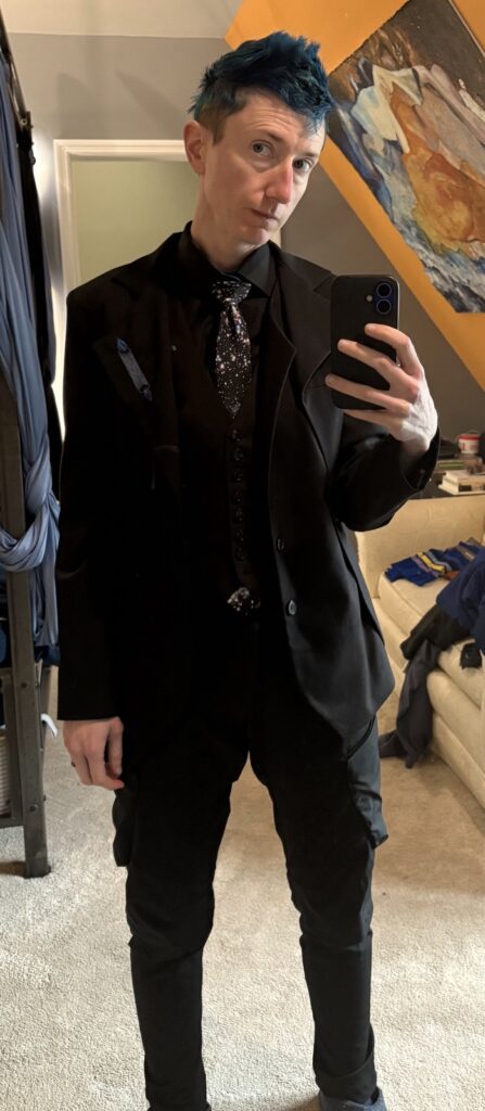 Willow in their bedroom wearing future tech black cargo pants, an asymmetrical tuxedo jacket, a many-buttoned black vest, black dress shirt, and a galaxy tie. The jacket has a blue candle burning at both ends embroidered on it.