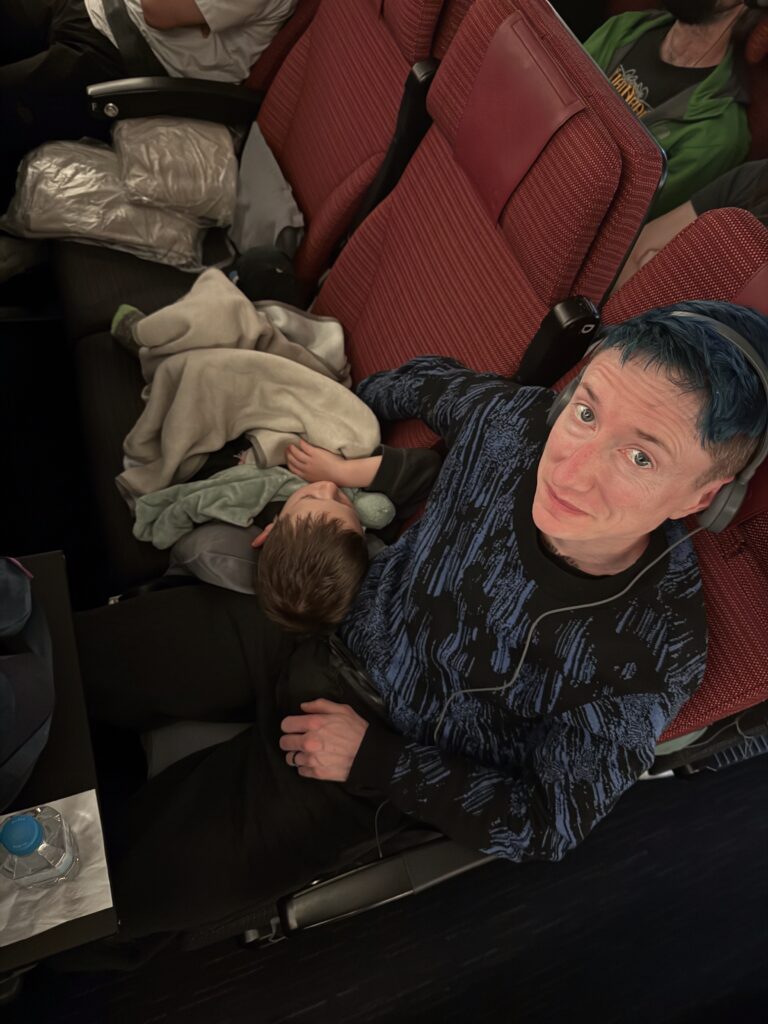 Willow looks up from their airplane seat while Locke sleeps with his head in their lap.